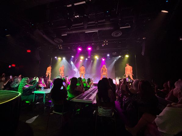 Packed nightclub cabaret: five male dancers on stage facing away, dropping underwear under colorful spotlights and haze while a seated audience watches and records on phones.