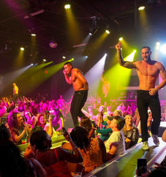 Shirtless male revue dancers performing on an illuminated runway in a packed nightclub, colorful stage lights and cheering audience snapping photos and clapping