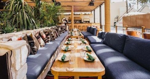 Sunlit outdoor patio dining with long wooden communal tables, navy cushioned benches lined with patterned throw pillows, lush greenery and neatly arranged place settings with green napkins under a pergola.