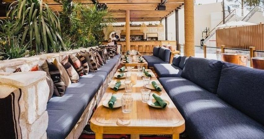 Sunlit outdoor patio dining with long wooden communal tables, navy cushioned benches lined with patterned throw pillows, lush greenery and neatly arranged place settings with green napkins under a pergola.