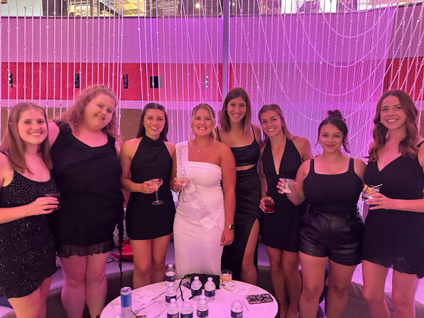Bachelorette party group photo in a pink-lit indoor lounge — women in black dresses and a bride-to-be in a white sash holding cocktails in front of a beaded curtain backdrop.