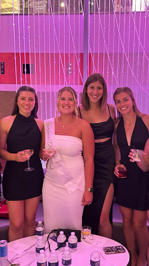 Bachelorette party group photo in a pink-lit indoor lounge — women in black dresses and a bride-to-be in a white sash holding cocktails in front of a beaded curtain backdrop.