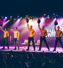 Five shirtless male performers in jeans tip black hats on a brightly lit stage during a live male revue, one singing into a microphone while audience silhouettes watch.