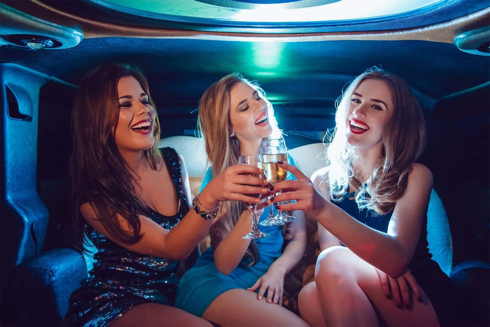 Three friends in evening dresses clinking champagne glasses inside a limousine with colorful party lighting for a night-out celebration.