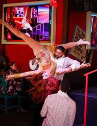 Drag performer in gold fringe costume and red wig flipped upside down mid-lift by a partner during a lively cabaret drag show in a colorful nightclub.