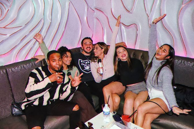 Six friends laughing and posing on a tufted sofa in an indoor karaoke lounge, holding microphones and drinks, arms raised against a wavy pink-lit wall for a celebratory night out.