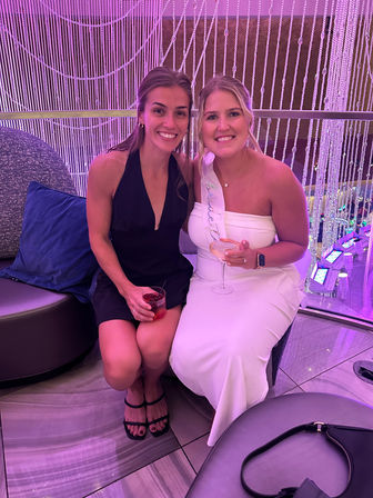 Two friends at a purple-lit cocktail lounge for a bachelorette party — bride-to-be in a white dress and sash holding a wine glass, companion in a black dress with a cocktail.