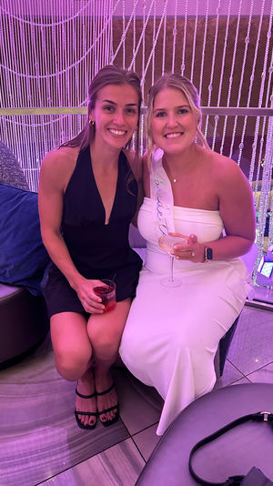 Two friends at a purple-lit cocktail lounge for a bachelorette party — bride-to-be in a white dress and sash holding a wine glass, companion in a black dress with a cocktail.