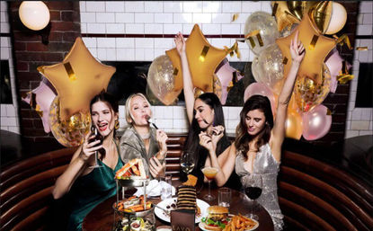 Four friends celebrating a festive night out in a restaurant booth with gold star and confetti balloons, cocktails, burgers, fries and a layered dessert tower.