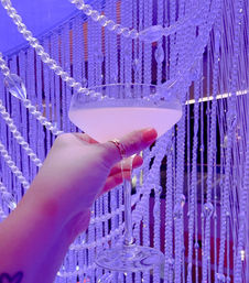 Hand with pink nails and rings holding a pale cocktail in a coupe glass against sparkling beaded curtains bathed in purple lounge lighting.
