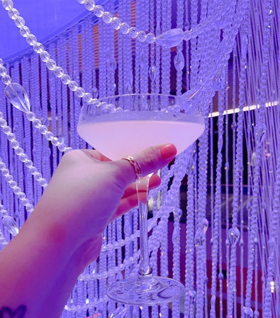 Hand with pink nails and rings holding a pale cocktail in a coupe glass against sparkling beaded curtains bathed in purple lounge lighting.