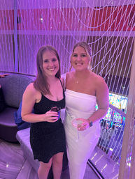 Two smiling women in cocktail dresses holding drinks in a purple-lit lounge with a beaded curtain backdrop and colorful slot machines below.