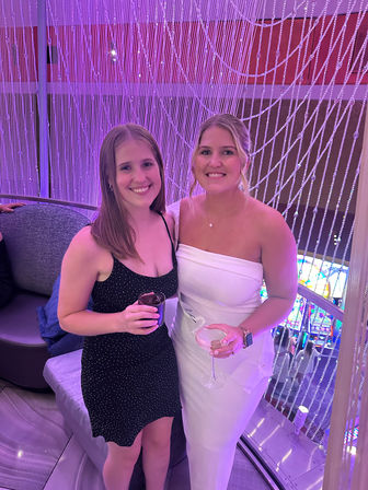 Two smiling women in cocktail dresses holding drinks in a purple-lit lounge with a beaded curtain backdrop and colorful slot machines below.
