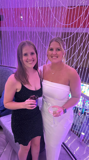 Two smiling women in cocktail dresses holding drinks in a purple-lit lounge with a beaded curtain backdrop and colorful slot machines below.
