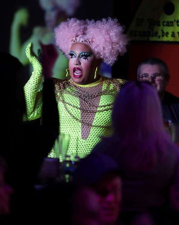 Drag performer in a pink curly wig and neon-green sequined costume with dramatic makeup and star earrings, singing onstage during a crowded nightclub drag show