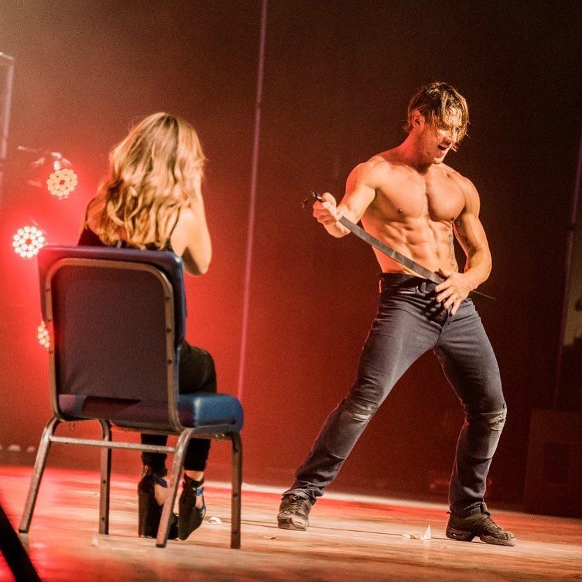 Shirtless muscular male performer in jeans holding a belt in a dramatic pose on a red-lit stage, entertaining a seated woman in high heels during a live cabaret-style dance performance.