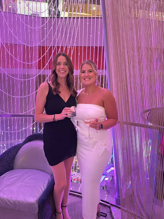 Two smiling women in cocktail dresses holding drinks in a pink-purple lit lounge, standing in front of shimmering beaded curtains at an indoor event