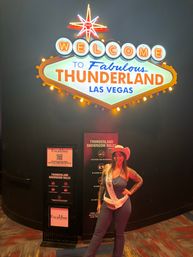 Bright retro Las Vegas-style neon "Welcome to Fabulous" marquee with starburst; woman in a cowboy hat and sash posing beneath at a showroom entrance.