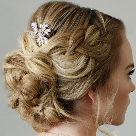 Blonde bridal updo with a braided crown leading into a textured low bun, loose face tendrils and a sparkly rhinestone hairpin, side profile