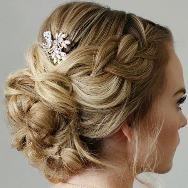 Blonde bridal updo with a braided crown leading into a textured low bun, loose face tendrils and a sparkly rhinestone hairpin, side profile