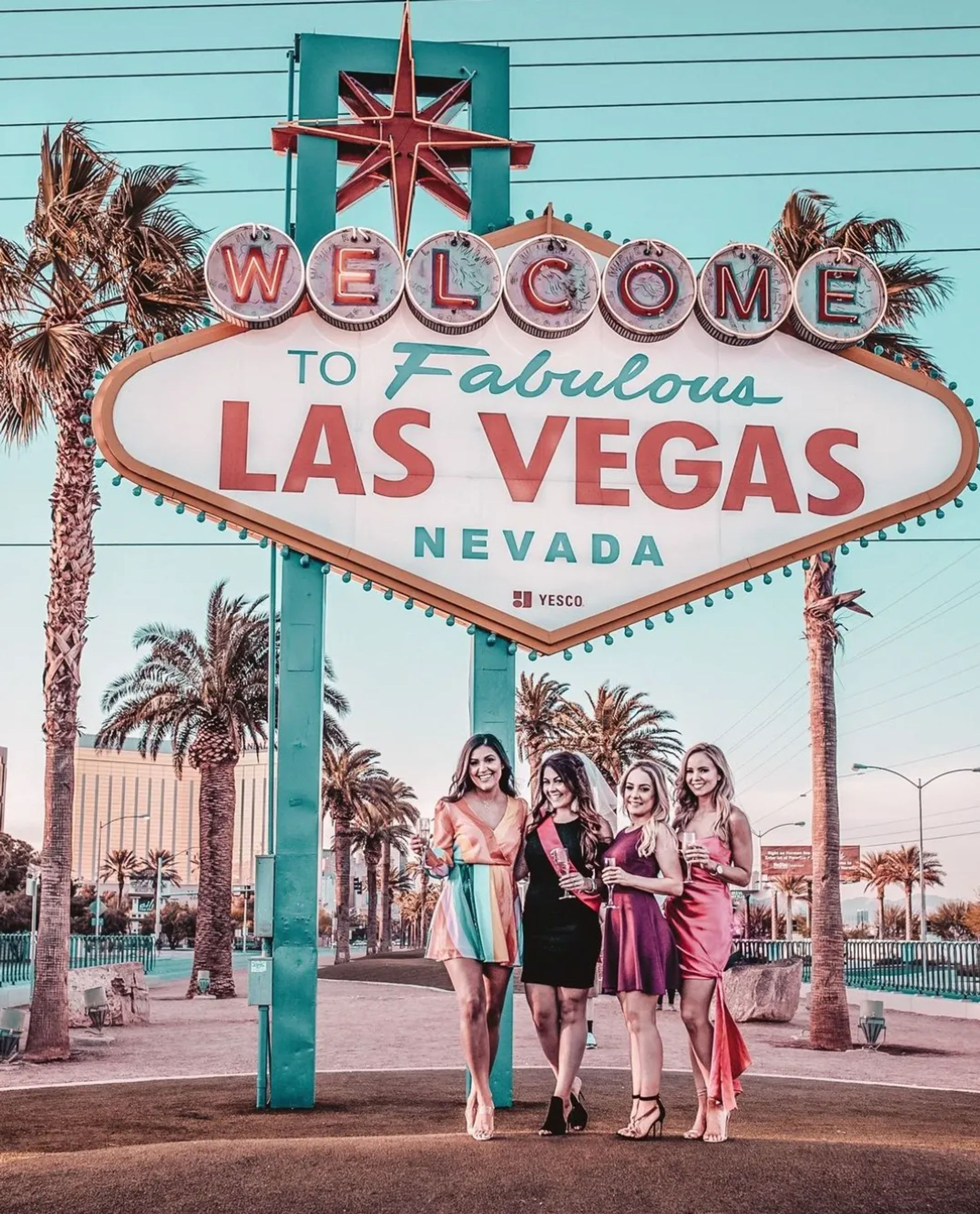 Four women in colorful party dresses posing beneath the iconic "Welcome to Fabulous Las Vegas Nevada" sign with palm trees and a pastel sky backdrop.