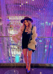 Stylish woman in a black mini dress, wide-brim hat and ruffled sheer sleeves posing by a glass railing in a purple-pink beaded LED light installation, wearing blue heels and a chain crossbody bag.