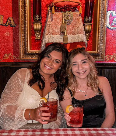 Two smiling friends seated at a cozy red-walled restaurant booth, each holding a lemon-garnished red cocktail over a red-and-white checkered tablecloth with ornate gold frame and tassel decor behind them.