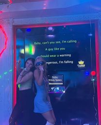 Two women singing into microphones in a neon-lit karaoke booth, night-out vibe with 'Toxic' lyrics on the screen