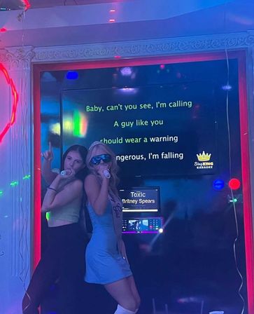 Two women singing into microphones in a neon-lit karaoke booth, night-out vibe with 'Toxic' lyrics on the screen
