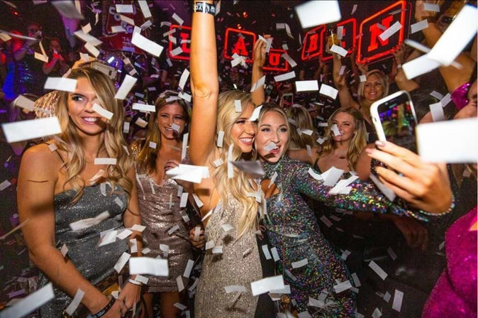 Group of women in glittery dresses taking a selfie as confetti rains down at a crowded nightclub with neon lights, lively party atmosphere