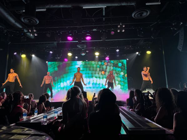 Shirtless male dancers performing a high-energy revue on a nightclub stage with colorful LED backdrop, purple and yellow spotlights, fog effects and an audience filming from table seating.