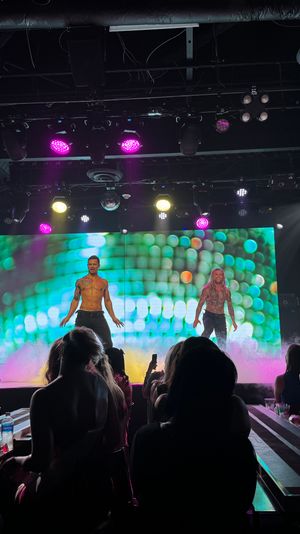 Shirtless male dancers performing a high-energy revue on a nightclub stage with colorful LED backdrop, purple and yellow spotlights, fog effects and an audience filming from table seating.