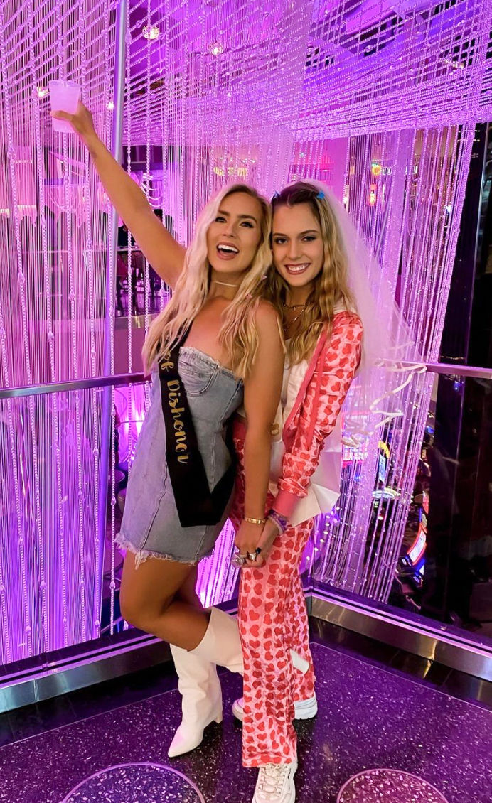 Two smiling women at a bachelorette party in a purple neon-lit lounge with beaded curtains, one wearing a sash and holding a drink.