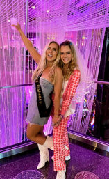 Two smiling women at a bachelorette party in a purple neon-lit lounge with beaded curtains, one wearing a sash and holding a drink.