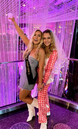 Two smiling women at a bachelorette party in a purple neon-lit lounge with beaded curtains, one wearing a sash and holding a drink.