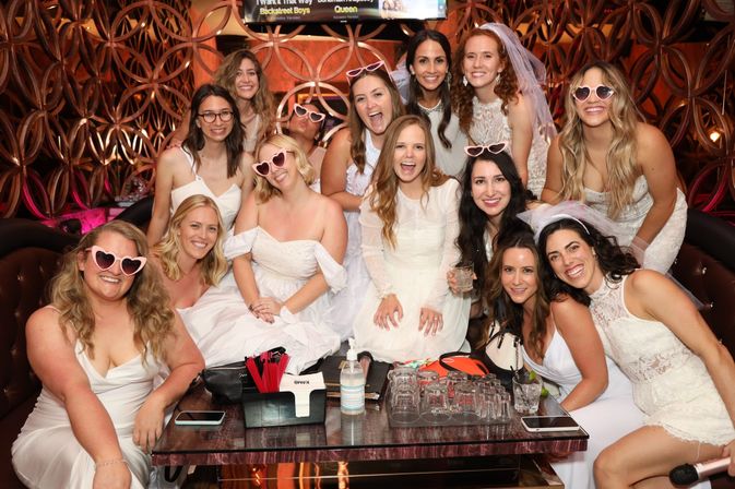 Bachelorette party group of women in white dresses, veils and heart-shaped sunglasses smiling in a stylish lounge booth with cocktails and glasses on the table