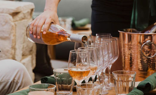 Hand pouring rosé wine into a row of wine glasses on a wooden patio table beside a copper ice bucket and folded green napkins.