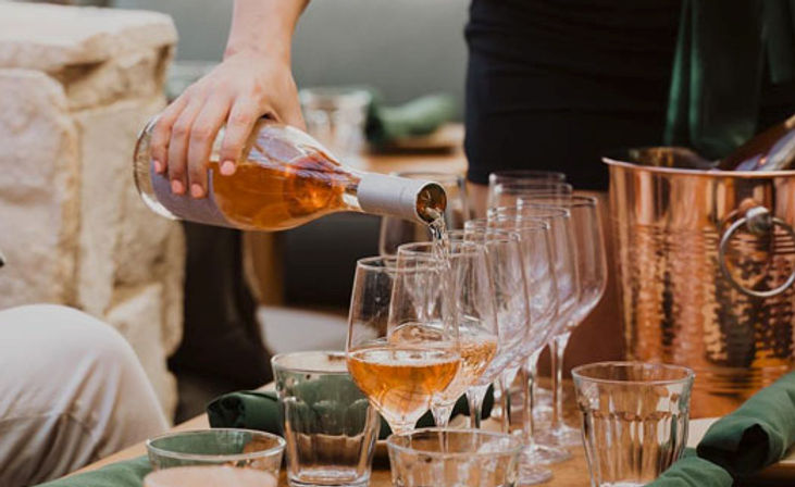 Hand pouring rosé wine into a row of wine glasses on a wooden patio table beside a copper ice bucket and folded green napkins.