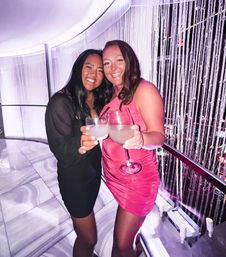 Two smiling friends in a pink dress and black dress clinking cocktail glasses in a purple-lit nightclub lounge with a beaded curtain and marble floor — celebratory night out.