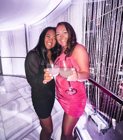 Two smiling friends in a pink dress and black dress clinking cocktail glasses in a purple-lit nightclub lounge with a beaded curtain and marble floor — celebratory night out.