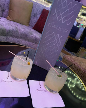 Two pale citrus cocktails in short glasses with pink and black straws on napkins, beside a tall purple menu on a glossy table in a plush, upscale cocktail lounge with velvet seating.