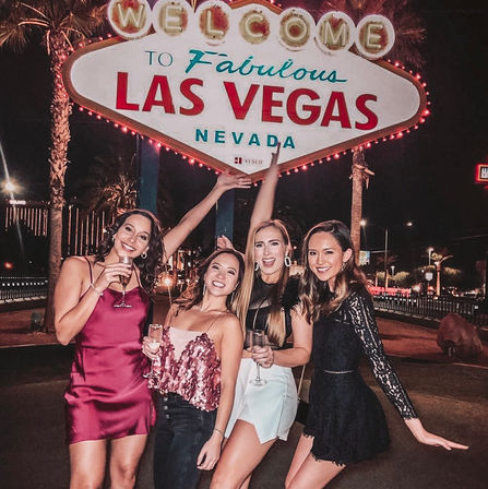 Four women in party outfits holding champagne, posing at night beneath the iconic ‘Welcome to Fabulous Las Vegas Nevada’ sign with palm trees and city lights