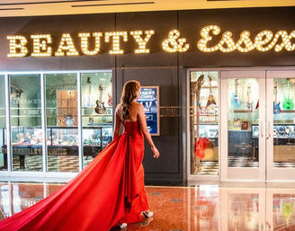 Back view of a person in a dramatic red evening gown with a long train walking across a glossy hotel-casino corridor past an illuminated marquee and glass display windows showing guitars and jewelry.