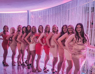 Glam group of women in pink, red and blush cocktail dresses posing in a line with hands on hips in front of a sparkling beaded curtain under pink neon lighting — girls' night out at a trendy lounge