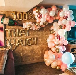 Indoor party backdrop with peach, pink, white and metallic gold balloon garland spilling from a giant champagne bottle balloon next to gold foil letter balloons on a patterned wall