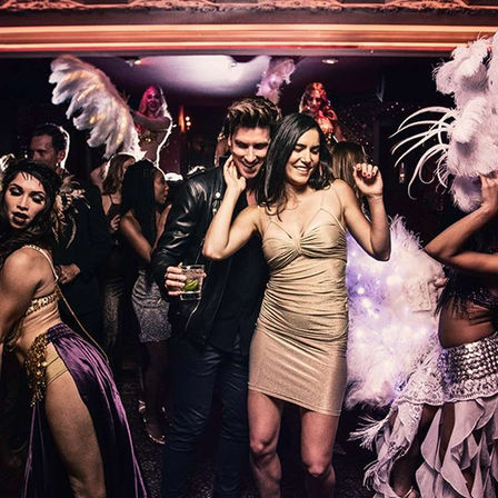 Couple dancing on a crowded nightclub dance floor, woman in a gold mini dress and man holding a cocktail, surrounded by showgirls in feathered cabaret costumes and lively partygoers.