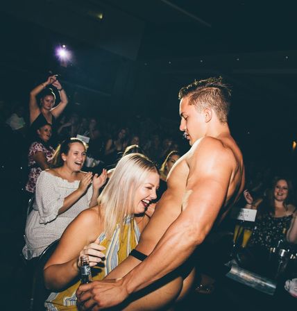 Bare-chested male dancer giving a playful lap dance to a laughing woman holding a drink in a crowded nightclub, audience cheering and clapping.