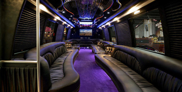 Luxury party bus interior with curved black leather bench seating, purple LED-lit aisle, mirrored ceiling chandelier, dance pole and widescreen TV with city lights visible through windows