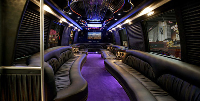 Luxury party bus interior with curved black leather bench seating, purple LED-lit aisle, mirrored ceiling chandelier, dance pole and widescreen TV with city lights visible through windows