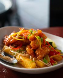 Crispy sweet-and-sour chicken with pineapple chunks, red and yellow bell peppers, bean sprouts and scallions, garnished with a lime wedge on a white restaurant plate